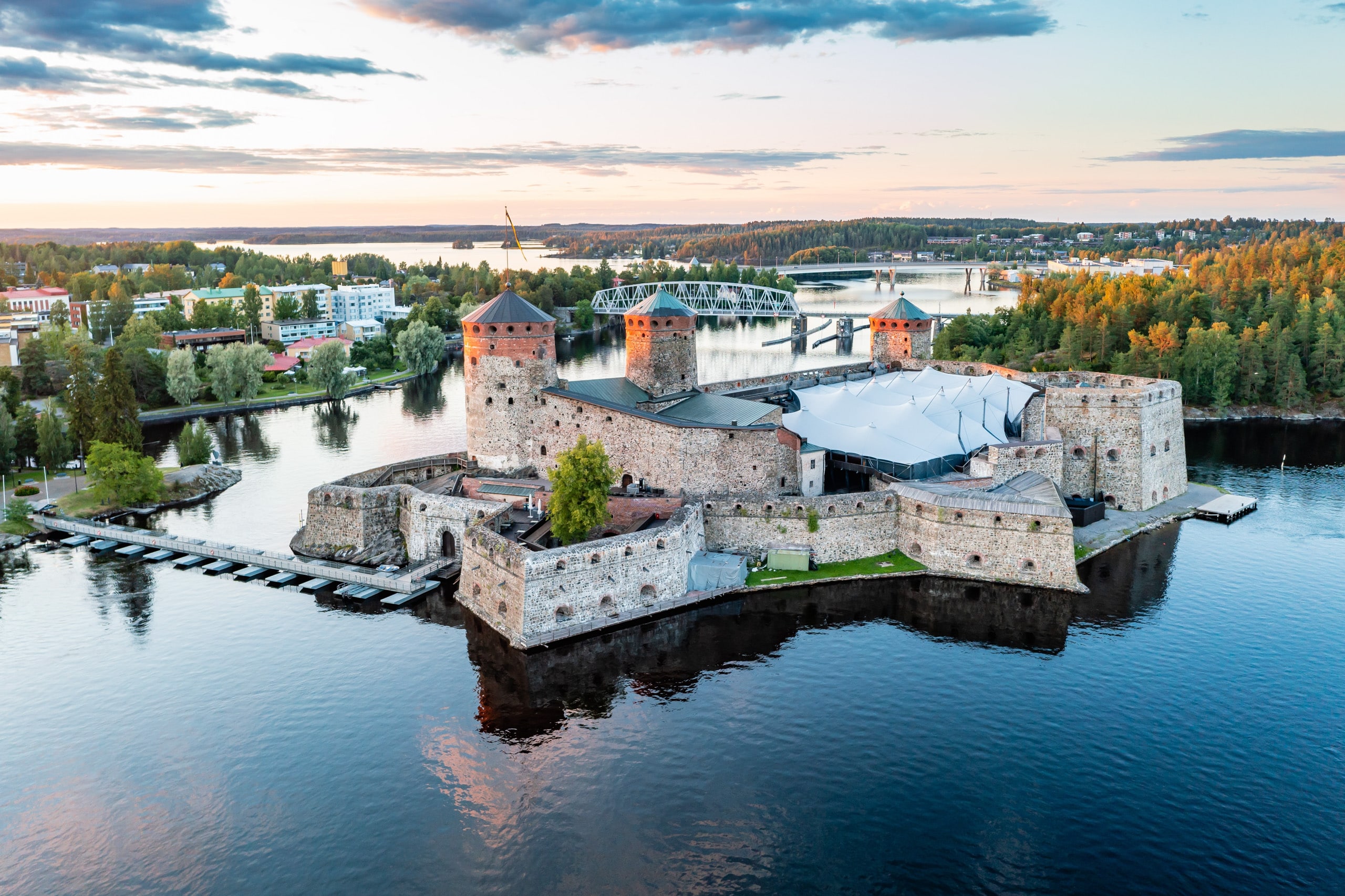 Ilmakuva Savonlinnan Olavinlinnasta ilta-auringossa, taustalla kaupunki ja siltoja Pihlajaveden yllä.